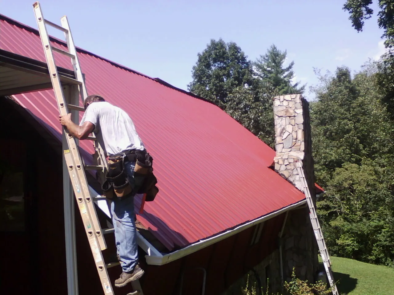 Roofer on a ladder beside a red metal roof and stone chimney — Soffit & Fascia Repair by Roofing Brothers in Merrimac